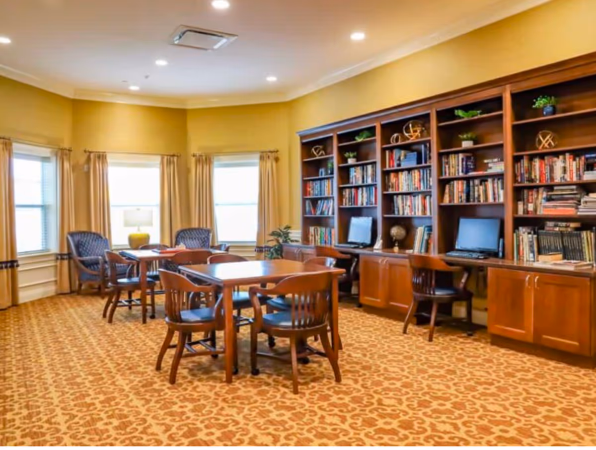 A cozy senior living common room with patterned carpet, wooden tables and chairs, a large built-in bookshelf filled with books and decorative items, and two computer workstations. The room has three windows with beige curtains and a table lamp between two armchairs.