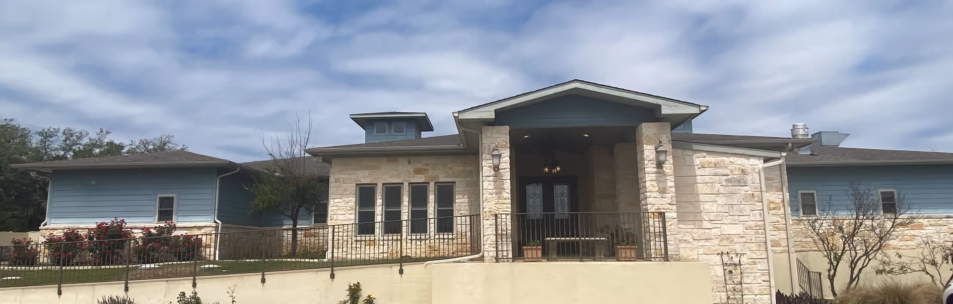 Front exterior of a single-story building with stone facade, blue siding, a covered central entrance and cloudy sky above.