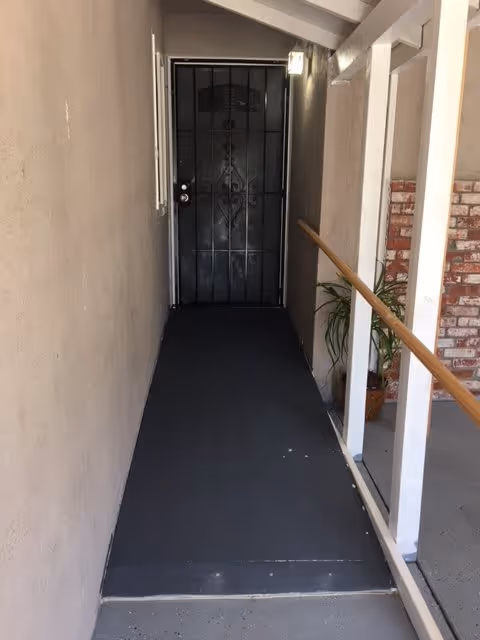 Narrow outdoor walkway with a black metal security door at the end. The walkway has a black floor and beige walls on the left side. On the right side, there is a white railing with a wooden handrail and a potted plant near the door. Part of a brick wall is visible on the right side beyond the railing.