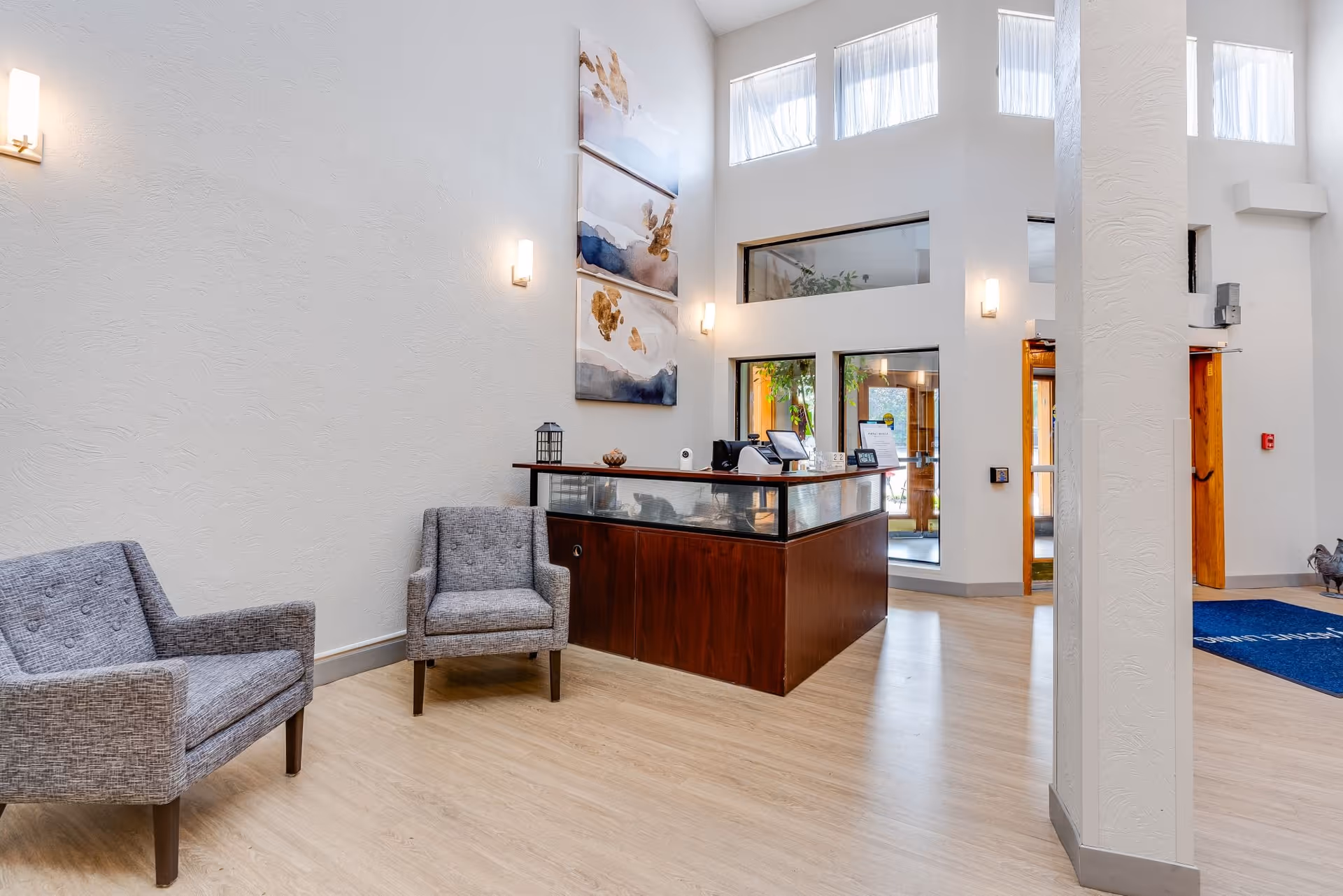 A bright and spacious reception area with two gray upholstered chairs on the left and a wooden reception desk with glass panels in the center. The walls are white with modern wall sconces and three abstract paintings hanging above the desk. Large windows near the ceiling allow natural light to fill the room, and a blue mat is visible near the entrance door on the right.