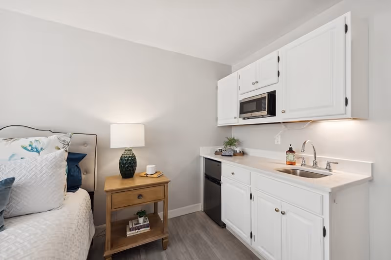 A small living space featuring a bed with multiple pillows on the left, a wooden nightstand with a lamp and a small plant, and a kitchenette on the right with white cabinets, a microwave, a mini fridge, a sink, and a soap dispenser.