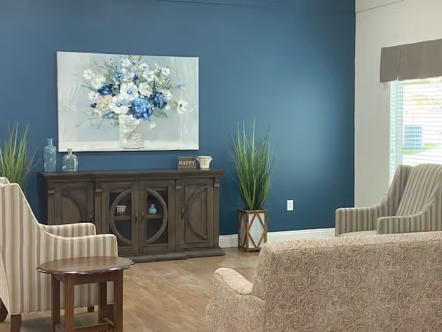 A cozy living room area with a dark wooden cabinet against a blue wall, decorated with a large floral painting, glass bottles, and a small 'HAPPY' sign. Two striped armchairs and a patterned sofa surround a small wooden side table. A tall green plant in a decorative pot is placed near the cabinet, and a window with blinds lets in natural light.