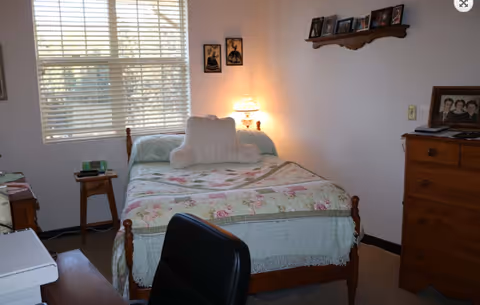 Small bedroom with a wooden bed covered in a floral quilt, a lit bedside lamp, dresser, and a window with blinds.