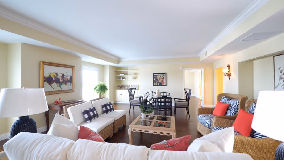 A bright and spacious living room with white and wicker furniture, including sofas and armchairs with red and black-and-white patterned cushions. A glass coffee table with decorative items is in the center. The room has wooden flooring, light yellow walls, a large window letting in natural light, and framed artwork on the walls. In the background, there is a dining area with a black dining table and chairs, and built-in shelves with decorative items.
