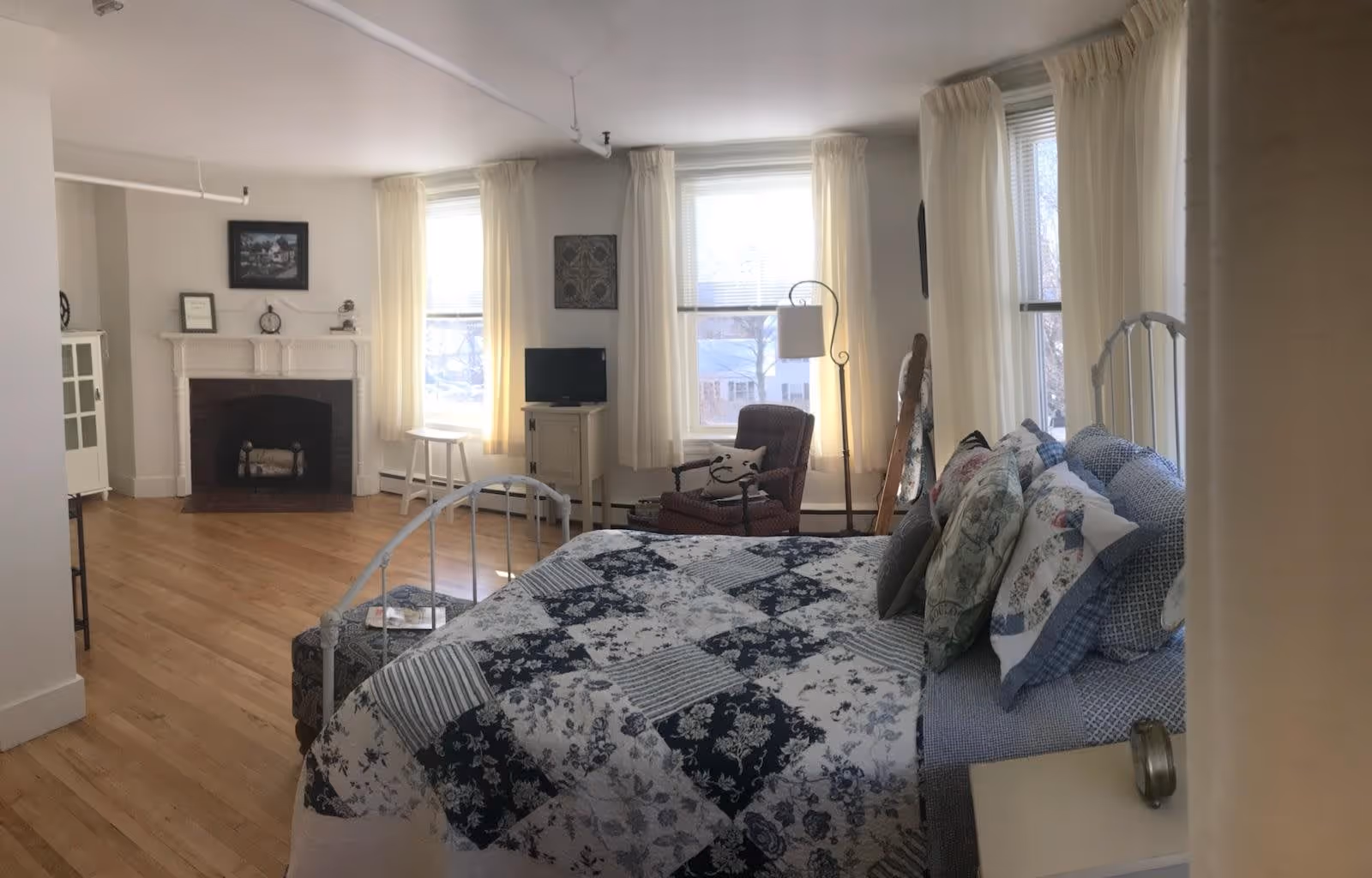 Sunlit bedroom with a metal bed topped with a patterned quilt, an armchair, fireplace, and large windows with sheer curtains.