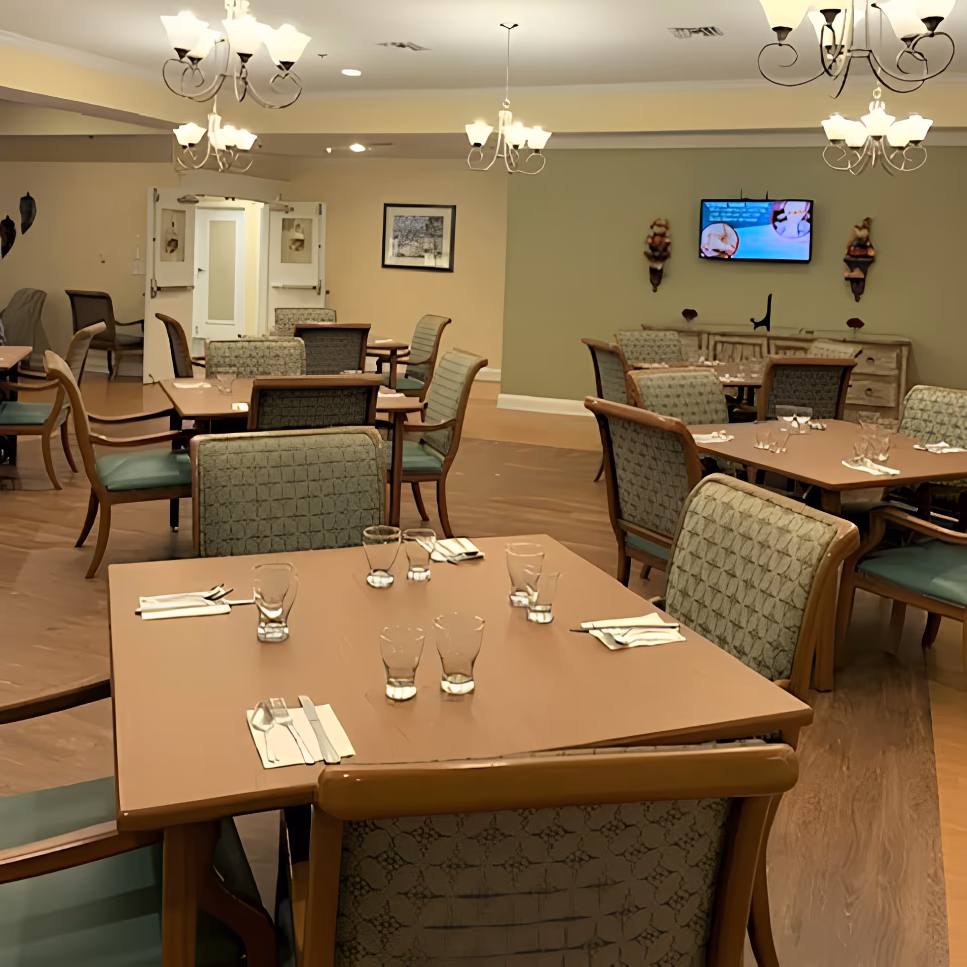 Empty dining room with multiple set tables, upholstered chairs, chandeliers, and a wall-mounted TV.