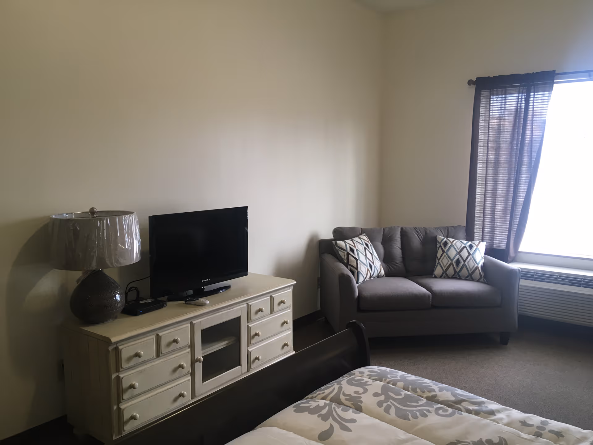 A small furnished bedroom showing a TV on a dresser, a lamp, a loveseat by a window with curtains, and part of a bed in the foreground.