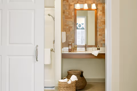 View into a bathroom featuring a white sliding door on the left, a shower with a silver showerhead, a vanity with a large mirror framed in wood, three light fixtures above the mirror, and two woven baskets on the floor holding towels.