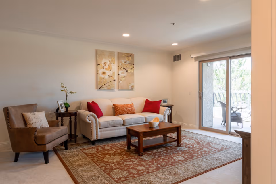 A cozy living room with a beige sofa adorned with red and orange cushions, a brown armchair with a decorative pillow, a wooden coffee table on a patterned area rug, two floral paintings on the wall, and a sliding glass door leading to an outdoor patio with chairs.