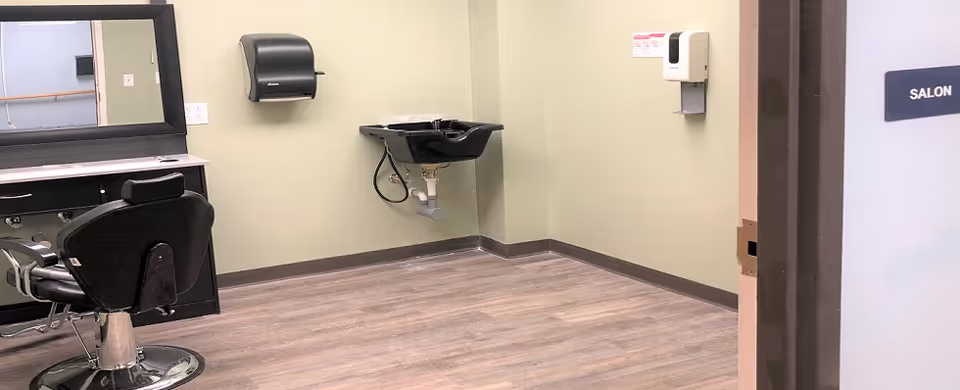 Empty salon room with a barber chair, mirror, wall-mounted hair-wash sink and dispensers.