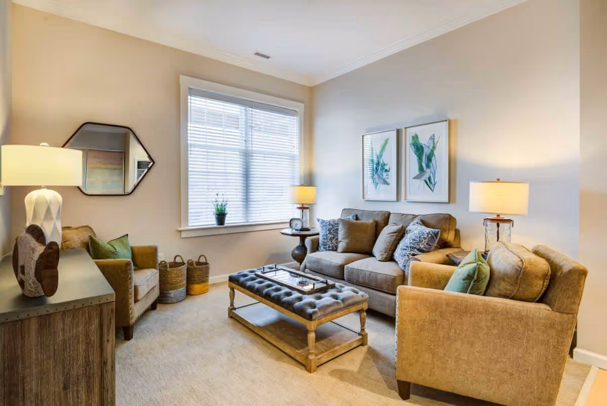 Sunlit living room with a sofa and two armchairs arranged around a tufted ottoman coffee table, lamps, wall art, and a window with blinds.