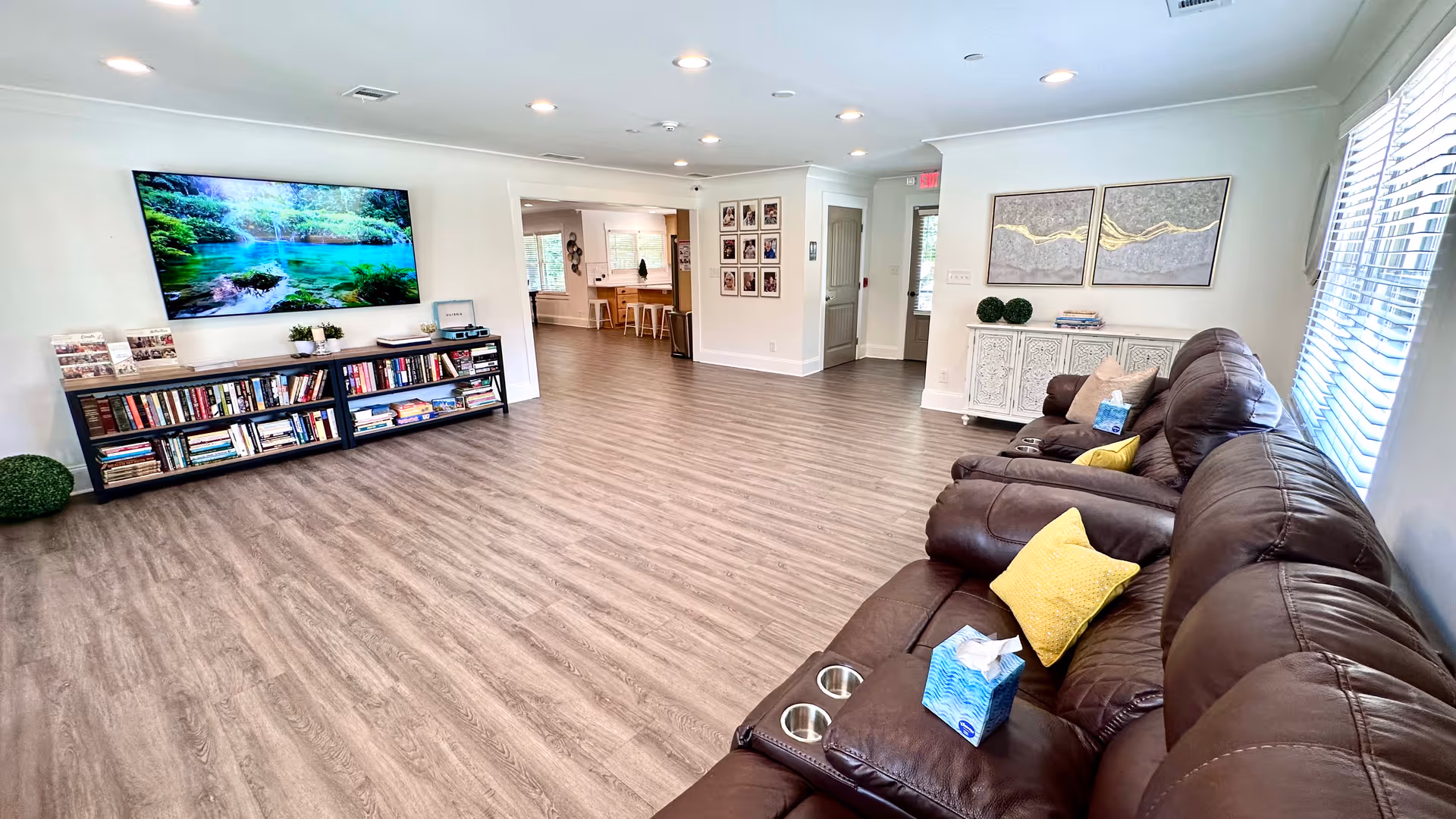 A spacious senior living common area with wood flooring, a large flat-screen TV mounted on the wall above a bookshelf filled with books, and a brown leather reclining sofa with yellow and beige pillows. The room has white walls, recessed lighting, large windows with blinds, and decorative artwork on the walls.