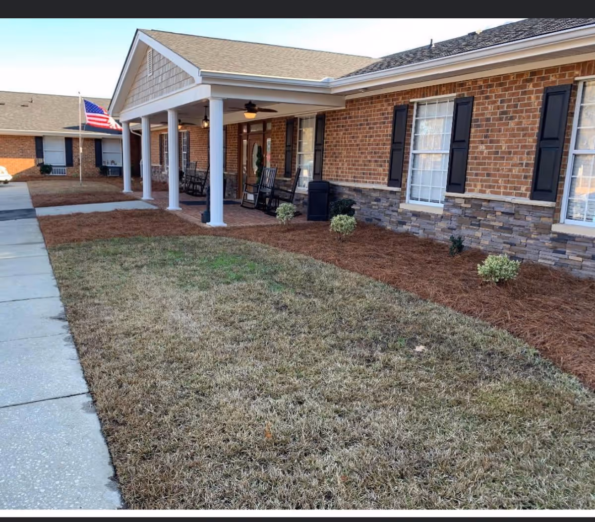 Front exterior of a brick building with a covered porch featuring rocking chairs, an American flag, and a landscaped lawn.