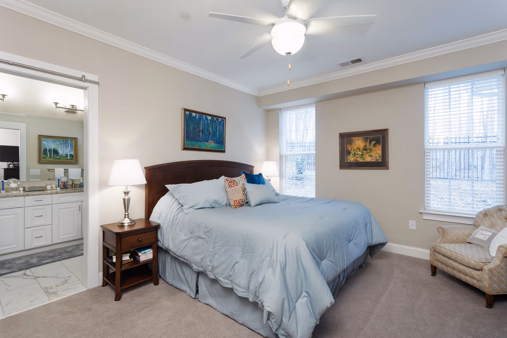 A cozy bedroom with a large bed covered in light blue bedding and several pillows. There is a wooden nightstand with a lamp and books next to the bed. Two windows with white blinds allow natural light into the room. A patterned armchair with a pillow is placed near the windows. The walls are decorated with two framed paintings. A ceiling fan with a light fixture is mounted on the ceiling. An open doorway leads to a bathroom with a granite countertop and white cabinetry.