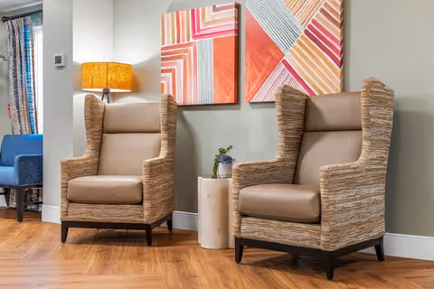 Two beige wingback chairs with wooden legs placed on a wooden floor next to a small cylindrical side table with a plant on it. Behind the chairs, there are two colorful abstract paintings on a light gray wall and a lit floor lamp with a yellow shade.