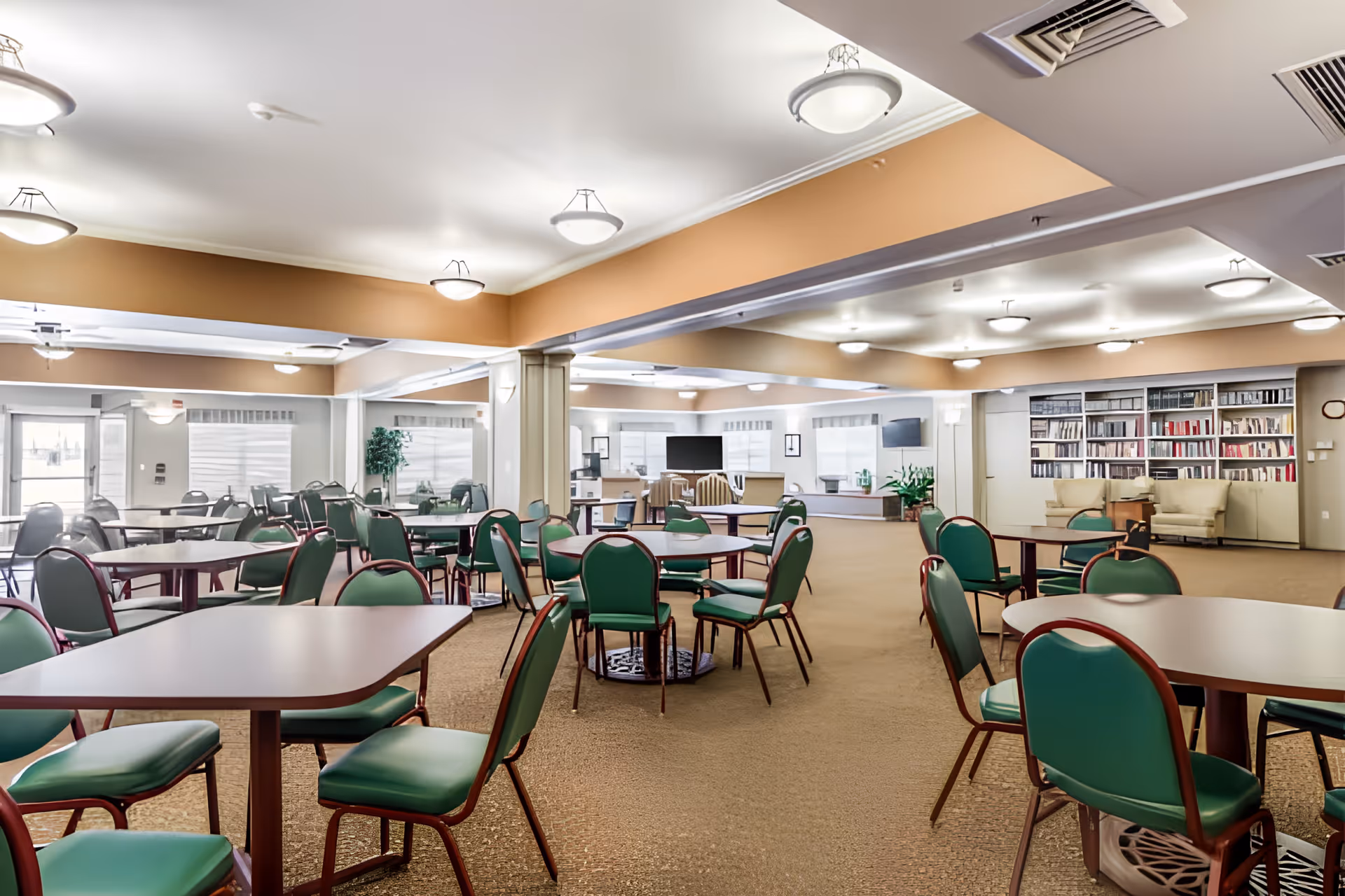 A spacious common area in a senior living facility with multiple tables and green cushioned chairs arranged throughout the room. The room features beige walls with a white ceiling and several ceiling lights. In the background, there is a seating area with armchairs, a large flat-screen TV, and a bookshelf filled with books. Large windows allow natural light to enter the space.