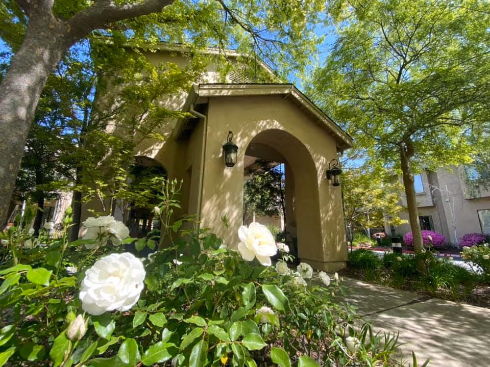 Arched entrance to a beige building framed by white roses, trees, and landscaped pathways.