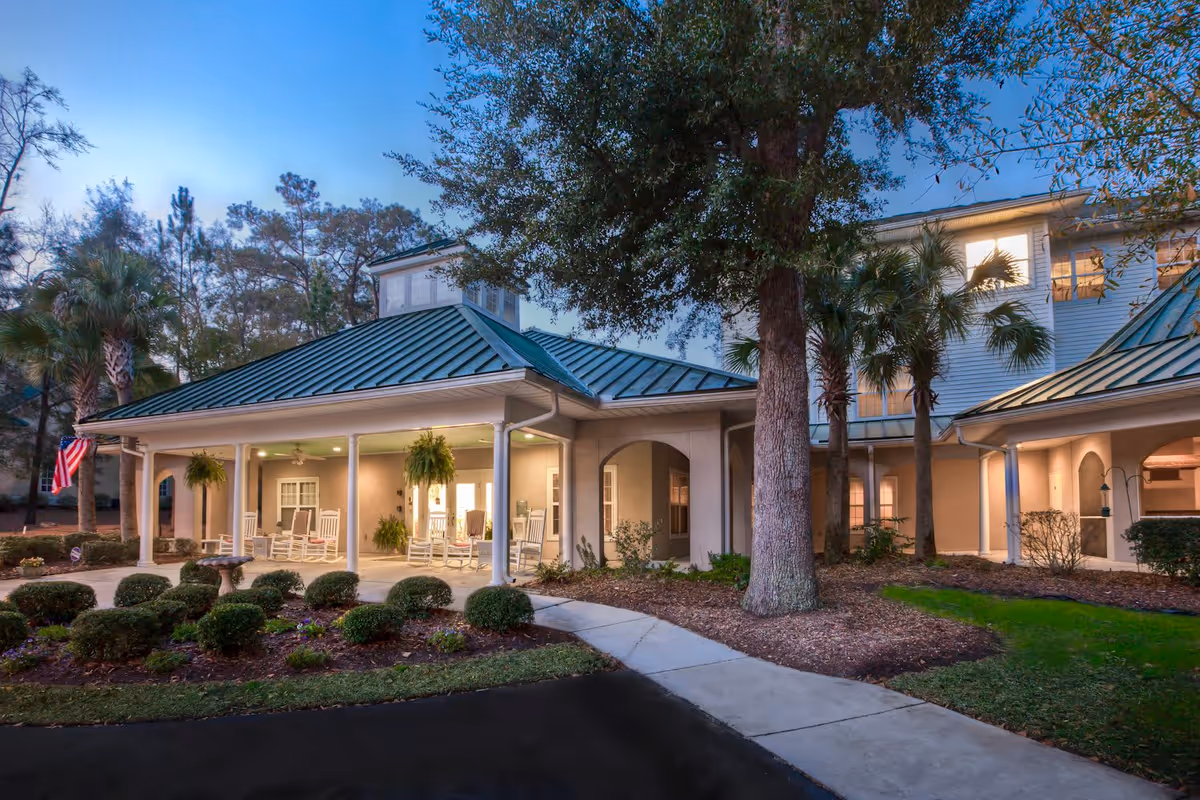 Entrance of a senior living facility with a covered porch, rocking chairs, hanging plants and landscaped shrubs under trees at dusk.