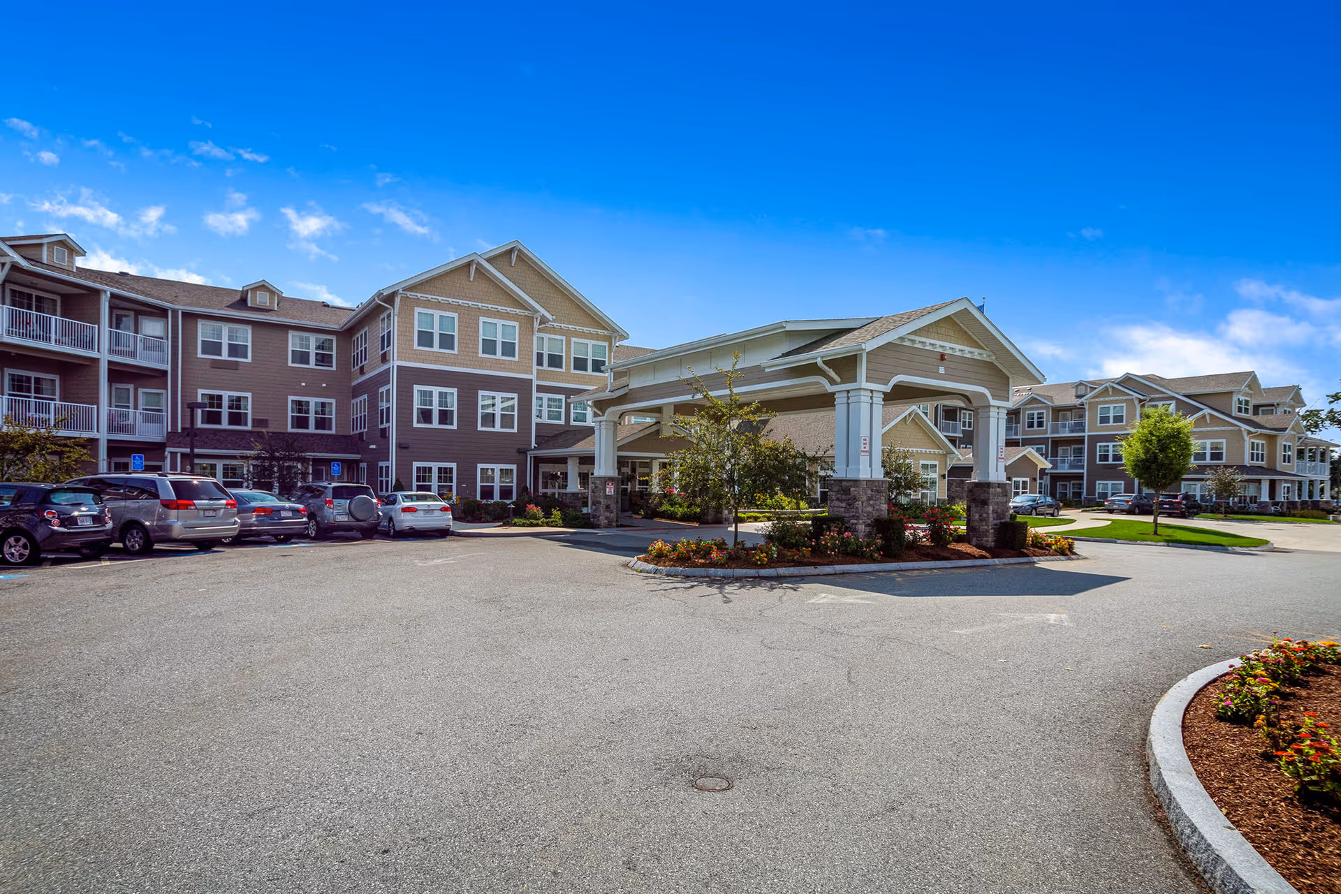 Exterior view of Fairview Estates Gracious Retirement Living facility showing a large three-story building with multiple windows and balconies. There is a covered entrance supported by stone pillars, surrounded by landscaped flower beds and small trees. Several cars are parked in the parking lot under a bright blue sky with scattered clouds.