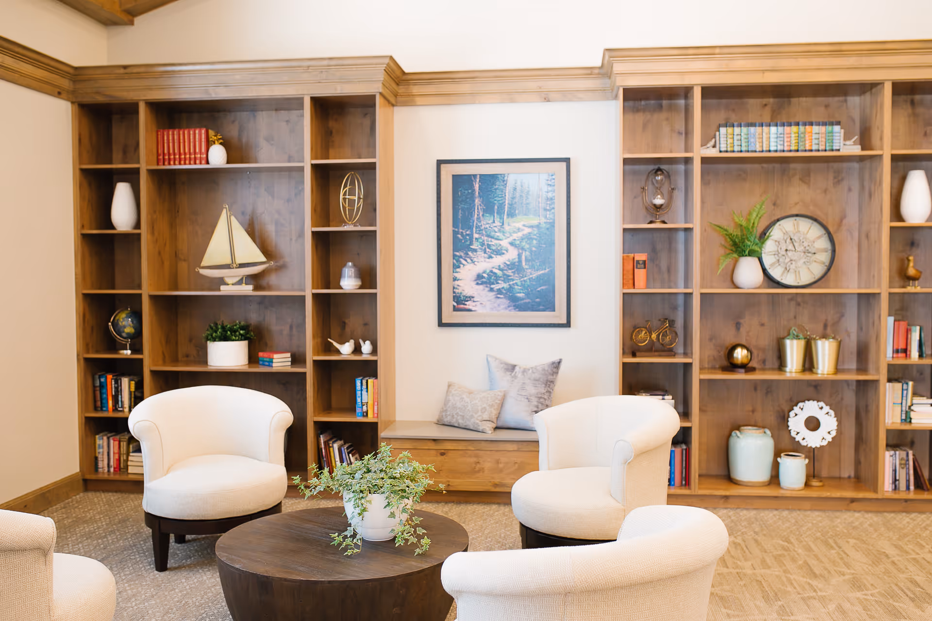 A cozy sitting area with four cream-colored armchairs arranged around a round wooden coffee table with a potted plant. Behind the chairs is a large wooden built-in bookshelf filled with books, decorative items, and plants. A framed picture of a forest path hangs on the wall in the center.