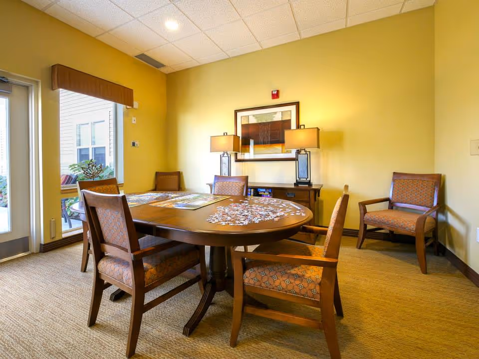 Small interior activity/dining room with a round wooden table surrounded by upholstered chairs, lamps, wall art, and puzzle pieces on the table.