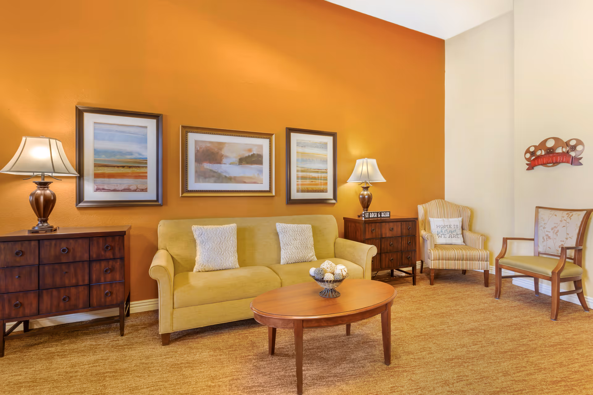 Cozy seating area with a yellow sofa, wooden coffee table, side cabinets and lamps against an orange accent wall with framed artwork.