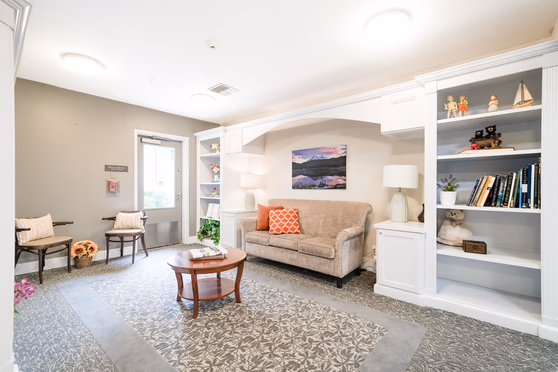 A bright and cozy sitting area in an assisted living facility featuring a beige couch with orange and patterned pillows, a round wooden coffee table with magazines, two wooden chairs with cushions, built-in white bookshelves with books and decorative items, a table lamp, and a door leading outside.