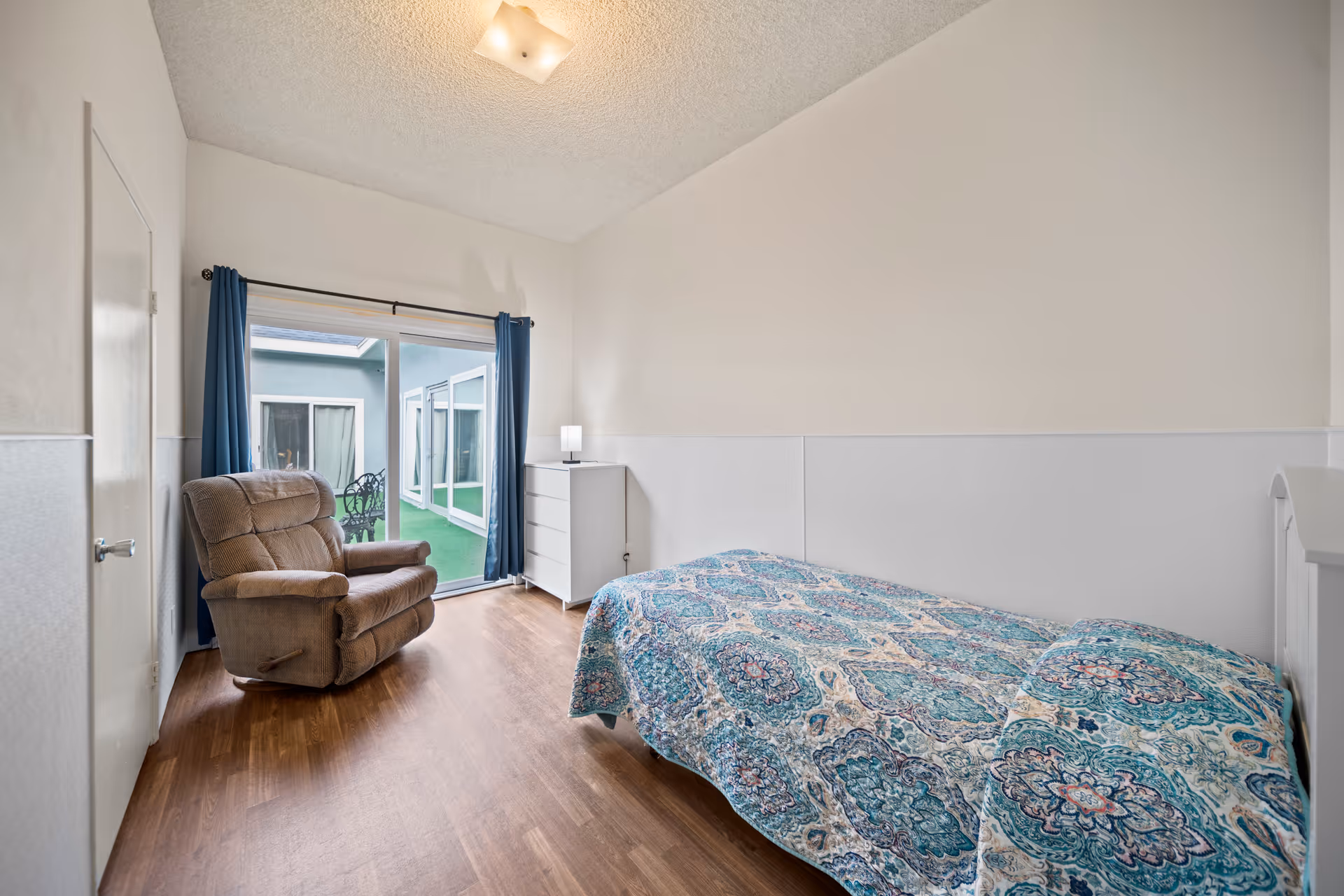 A simple bedroom with a single bed covered in a blue patterned quilt, a brown recliner chair, a white dresser with a small lamp on top, and a sliding glass door with blue curtains leading to an outdoor patio area.