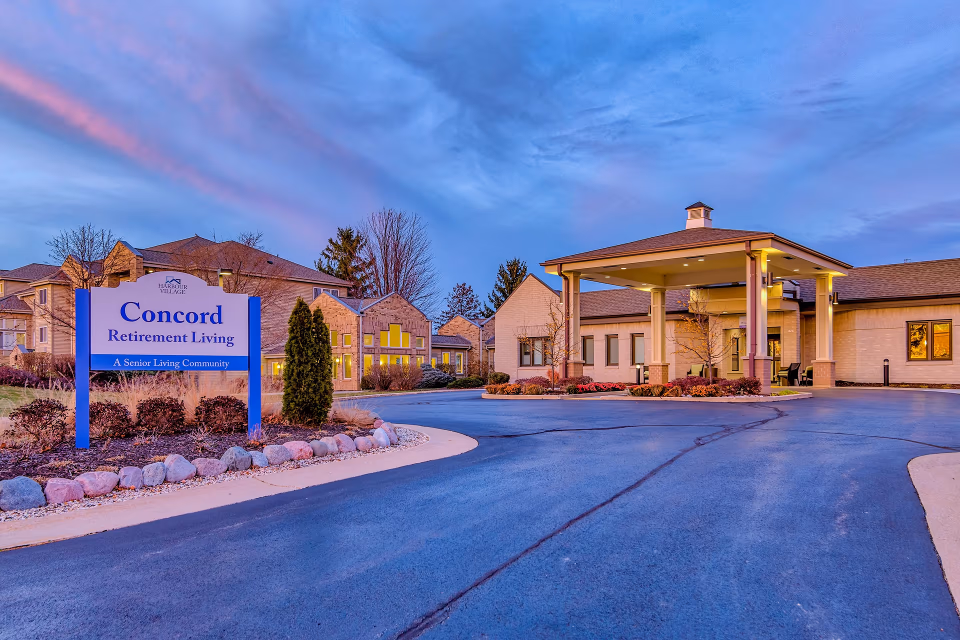 Exterior view of Concord Retirement Living, a senior living community, during dusk with a clear sky showing some clouds. The building has a covered entrance with columns and warm lighting. There is a sign in the foreground that reads 'Concord Retirement Living, A Senior Living Community' with landscaping around it.