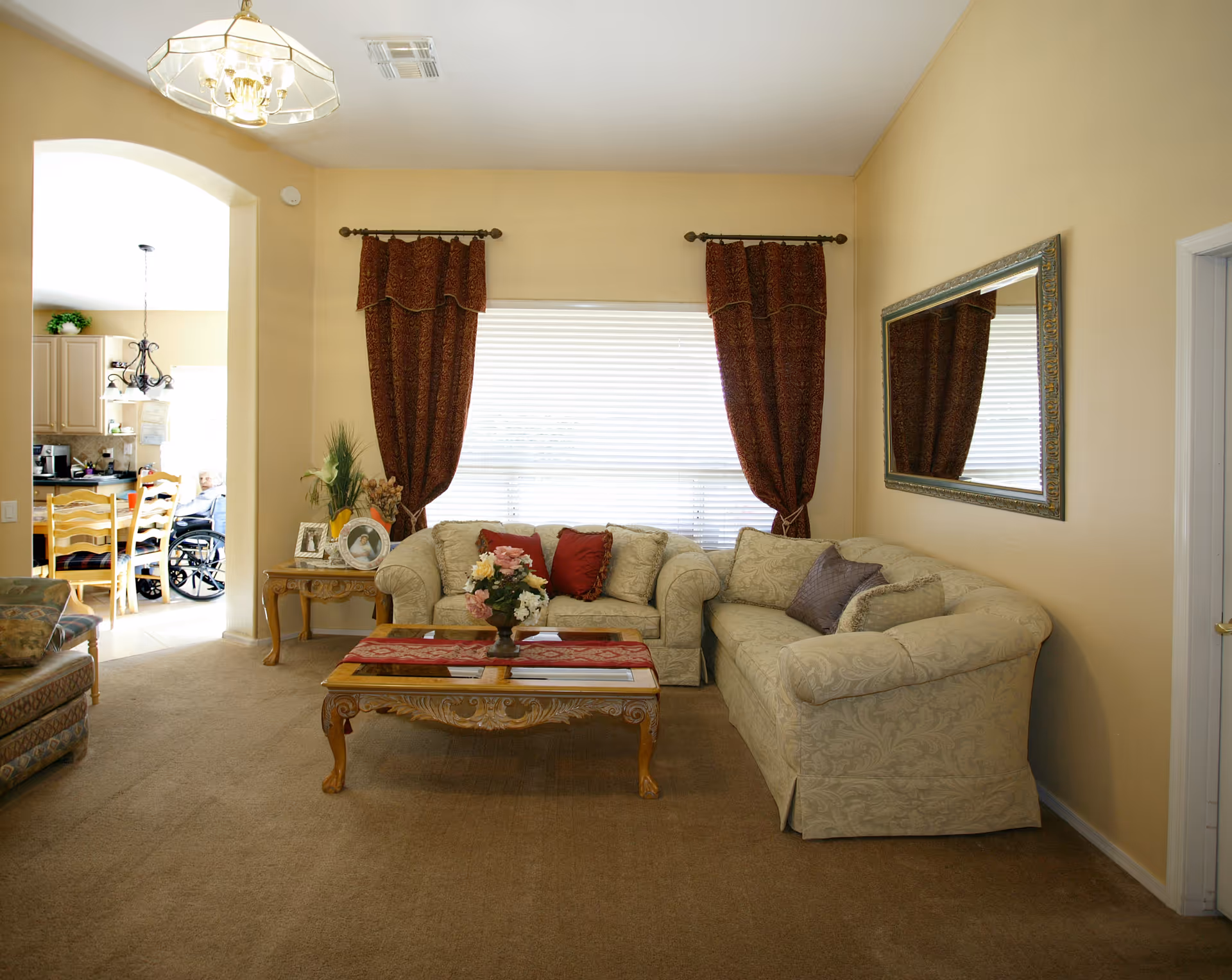 A cozy beige living room with patterned sofas, a wooden coffee table, and burgundy curtains; a kitchen area with people seated in the background.