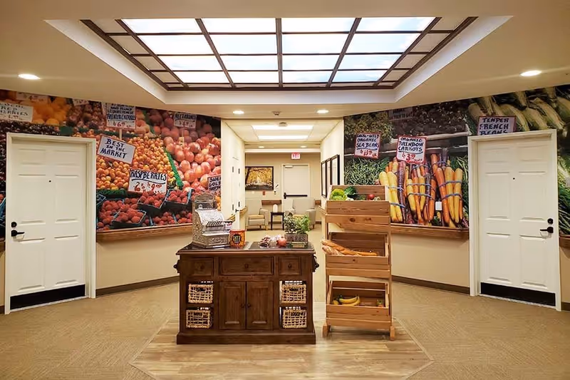 Interior hallway area with two white doors on either side and a wooden display table and wooden produce rack in the center. The walls are decorated with large murals of fresh fruits and vegetables with price signs, creating a market-like atmosphere. The ceiling features a large skylight with a grid pattern.