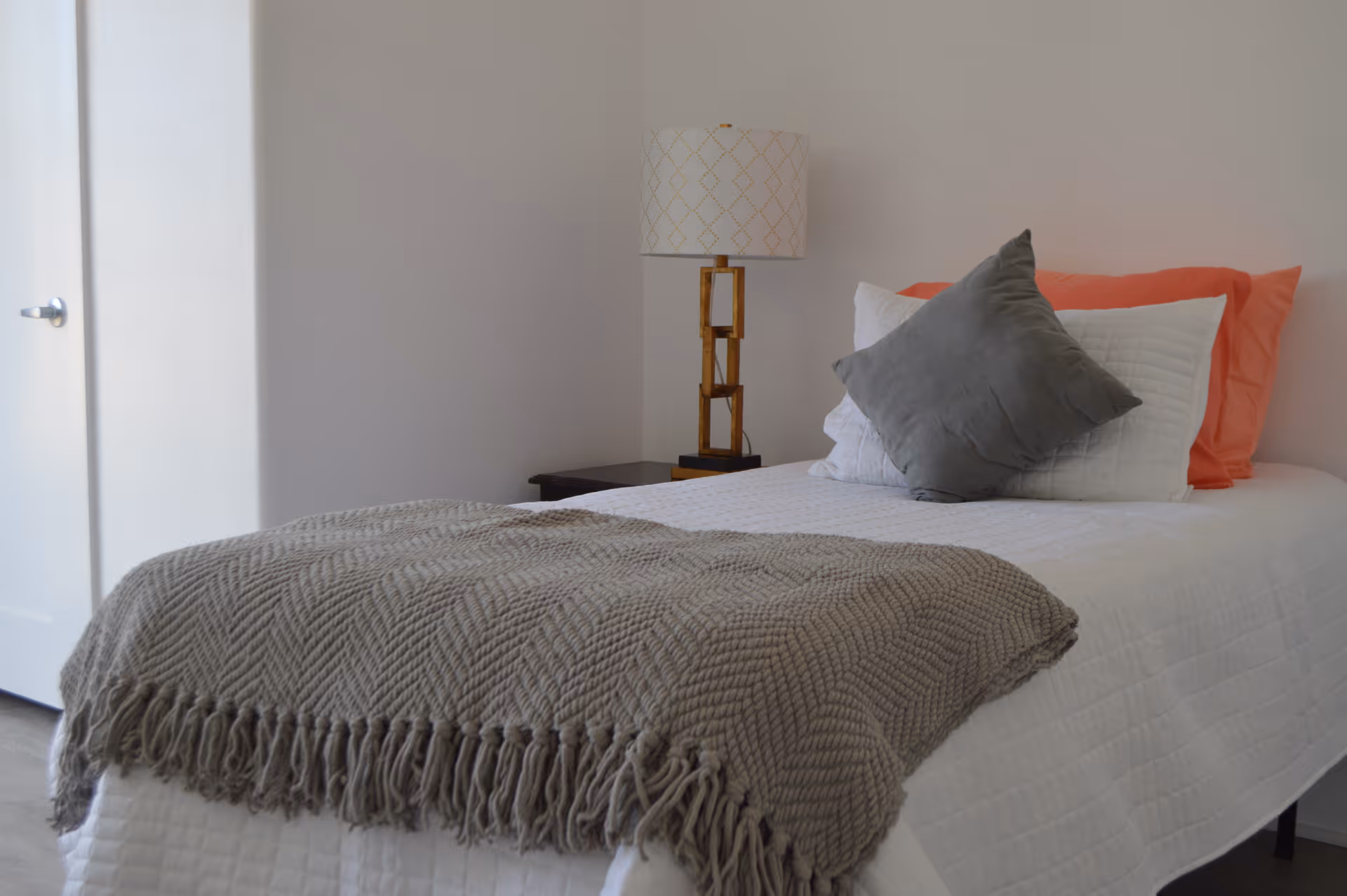 A neatly made single bed with white bedding, a gray throw blanket, coral and gray pillows, and a bedside lamp in a minimalist bedroom.