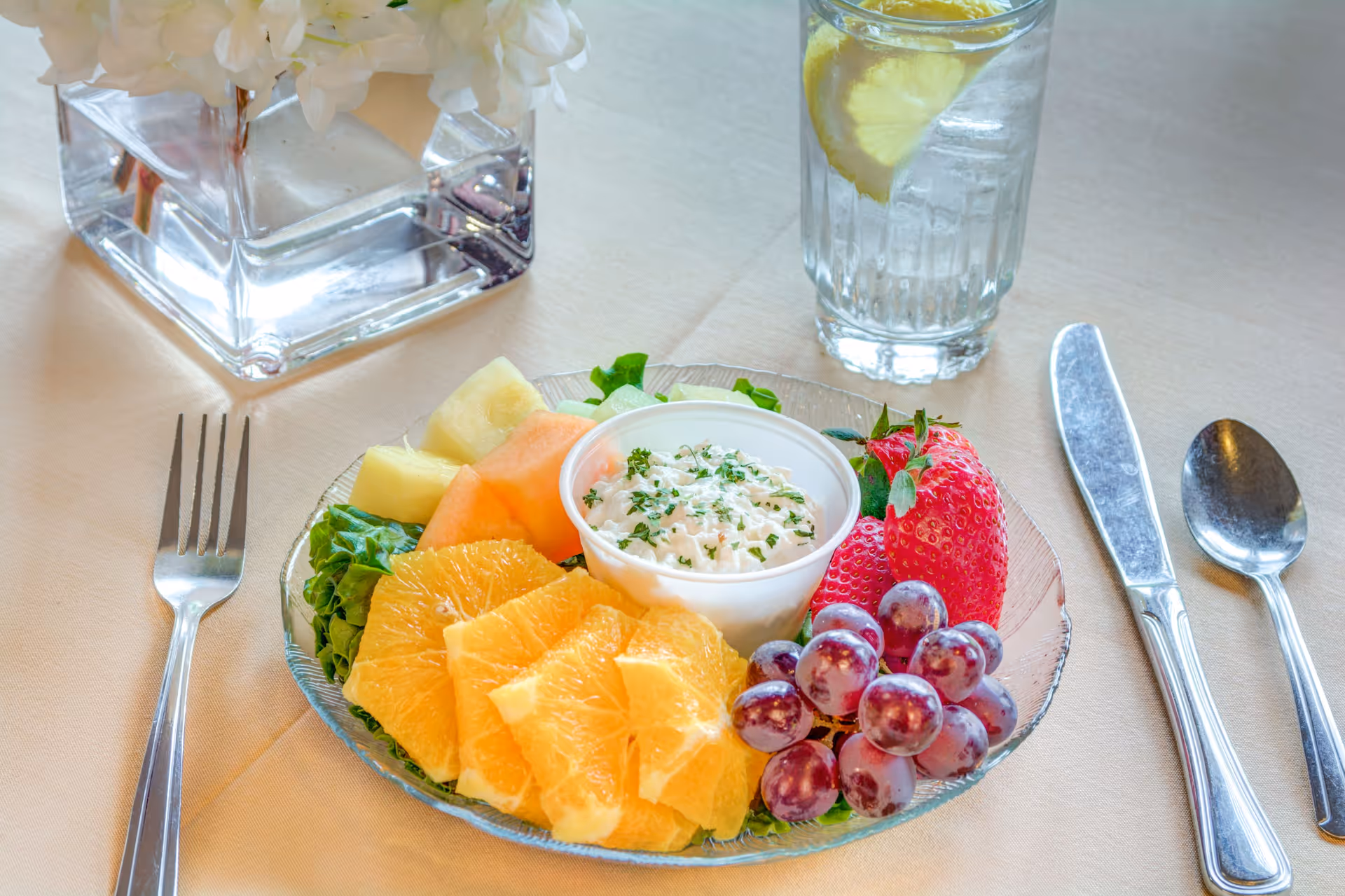A glass plate with a fresh fruit salad including orange slices, grapes, strawberries, cantaloupe, and pineapple, accompanied by a small cup of cottage cheese garnished with herbs. The plate is set on a beige tablecloth with a fork on the left and a knife and spoon on the right. A glass of water with a lemon slice and a square glass vase with white flowers are also visible on the table.