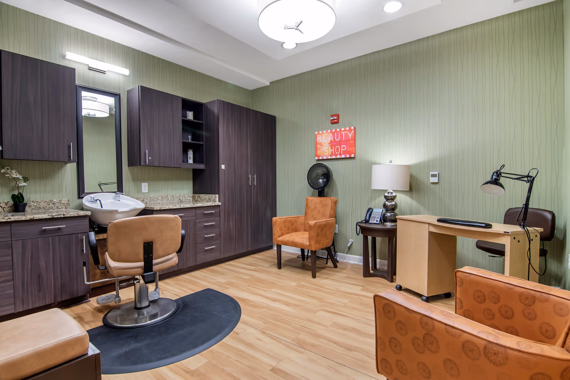 Interior of a beauty shop room with a salon chair in front of a sink and mirror, dark wood cabinets, two orange patterned armchairs, a small round table with a lamp and phone, a desk with a lamp and chair, and light wood flooring with green textured wallpaper.