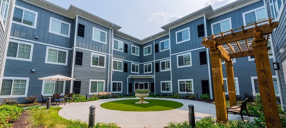 Three-story gray residential building surrounding a landscaped central courtyard with a circular walkway, fountain, seating, and a wooden pergola.