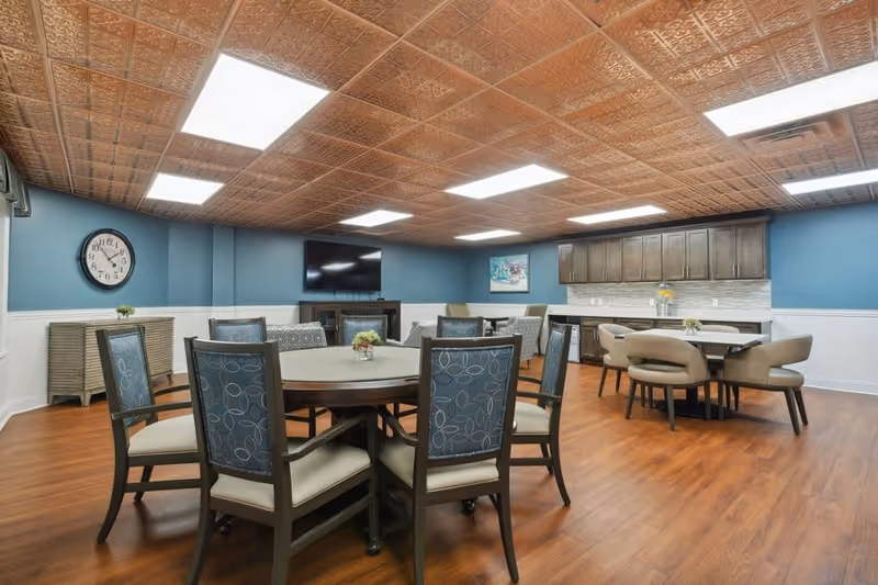 A spacious senior living common area with round tables and cushioned chairs arranged for dining or socializing. The room features wood flooring, blue walls with white wainscoting, a decorative copper-colored ceiling, and bright overhead lighting. There is a wall-mounted flat-screen TV, a sideboard with a clock above it, and a kitchenette area with cabinets and a countertop in the background.