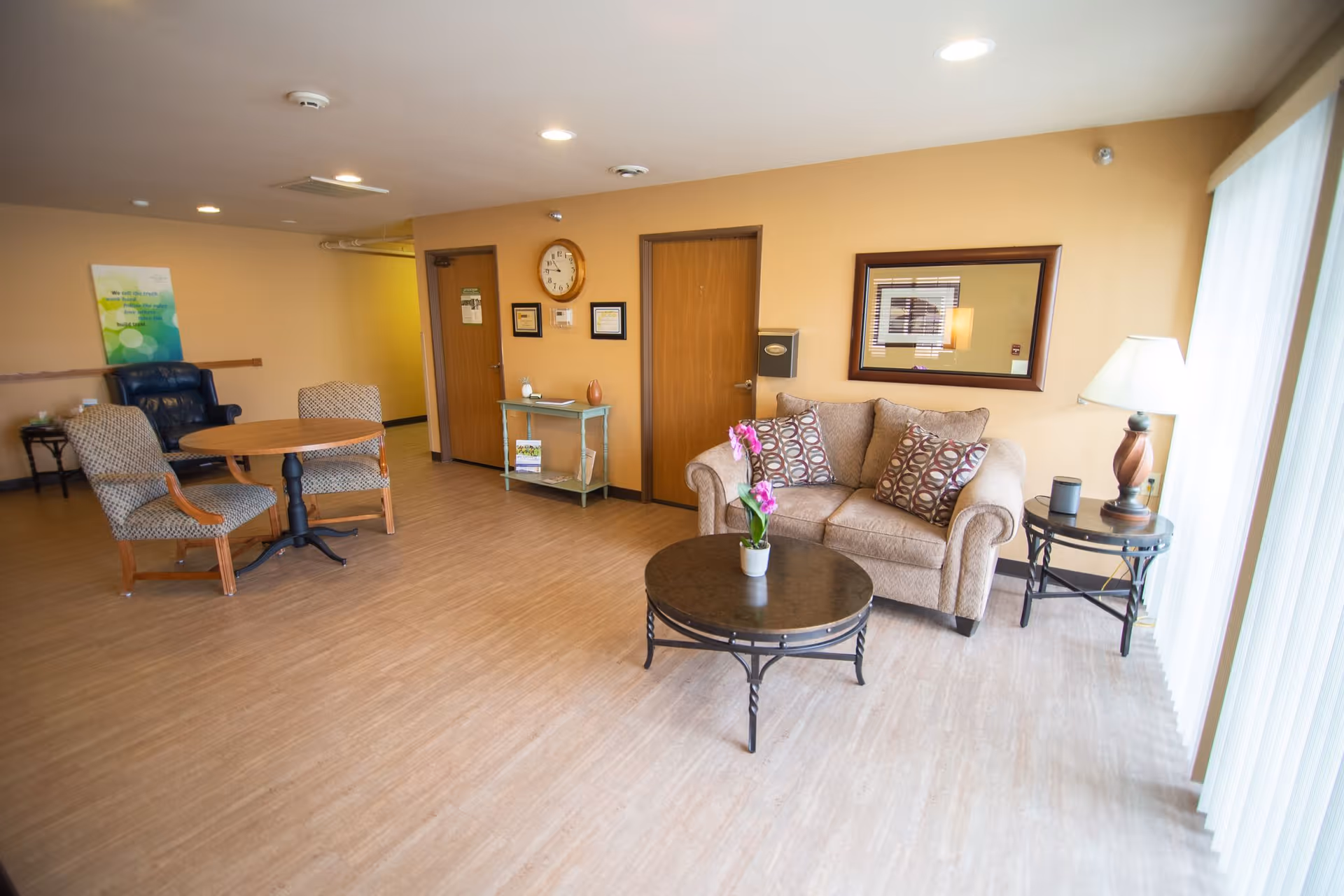 A cozy living room area with a beige loveseat adorned with patterned cushions, a round coffee table with a potted orchid, a side table with a lamp, and a round wooden table with two upholstered chairs. The room has light wood flooring, beige walls, two wooden doors, a wall clock, framed certificates, and a large window with vertical blinds allowing natural light to enter.