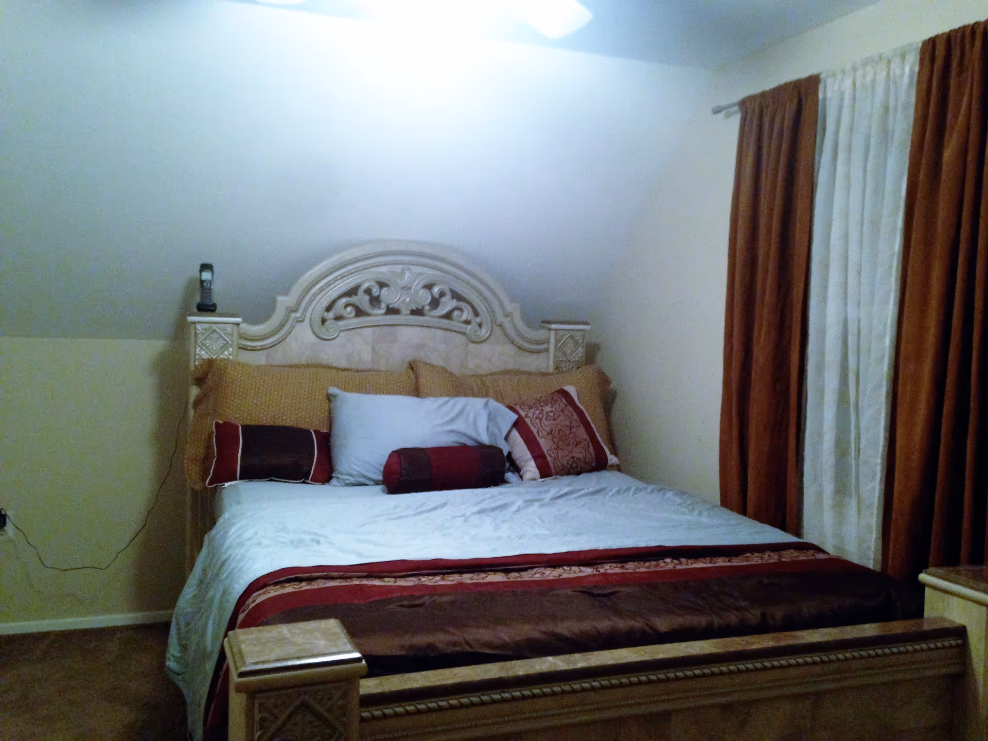 A bedroom with a large ornate wooden bed featuring a carved headboard. The bed is made with white sheets, a brown and red blanket, and several pillows in shades of brown, red, and white. To the right, there are brown and cream curtains covering a window. The walls are light-colored, and there is a phone placed on the left side of the headboard.