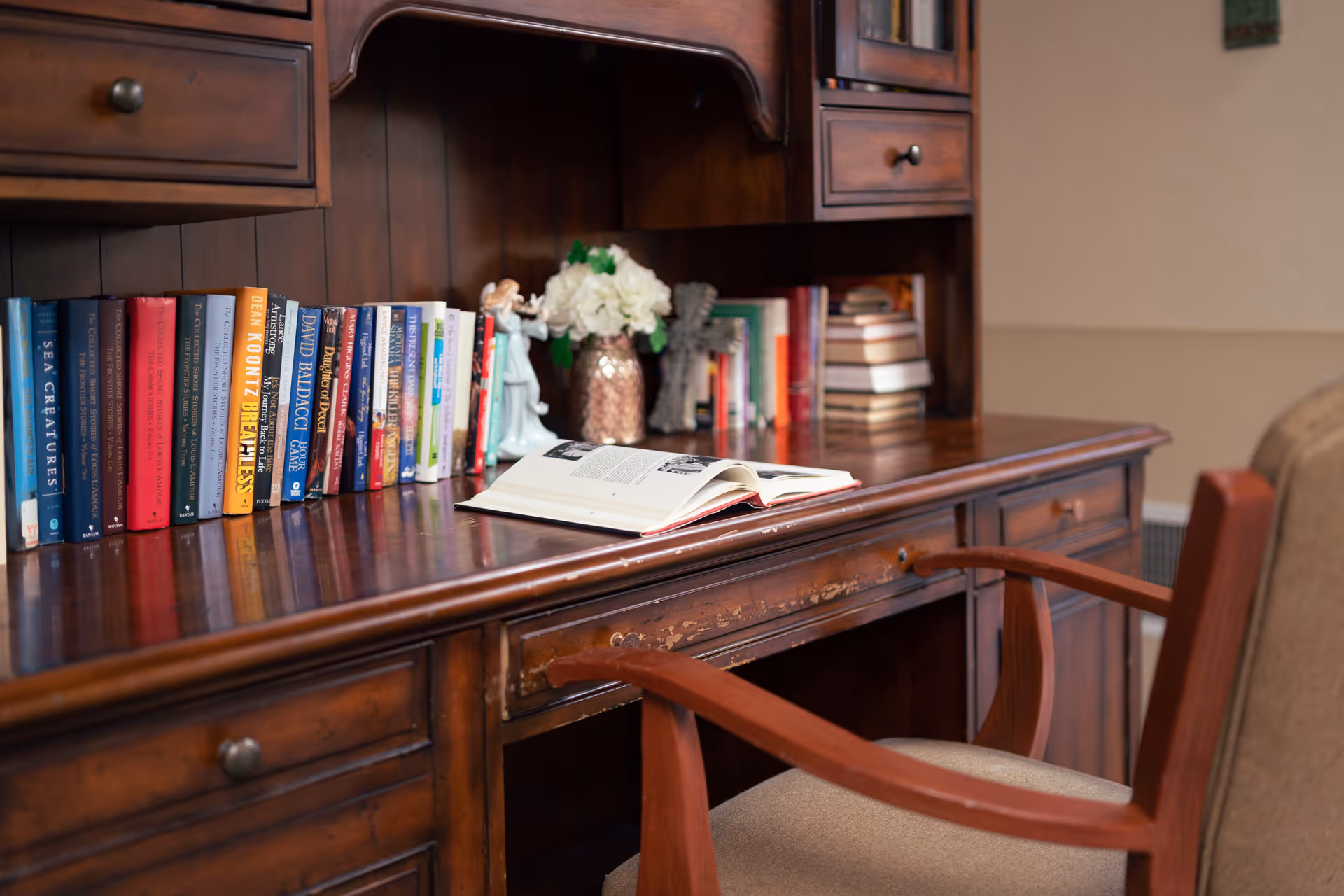 A wooden desk with multiple drawers and a hutch filled with books, a vase of white flowers, and decorative figurines. An open book lies on the desk surface, and a wooden chair with beige upholstery is positioned in front of the desk.