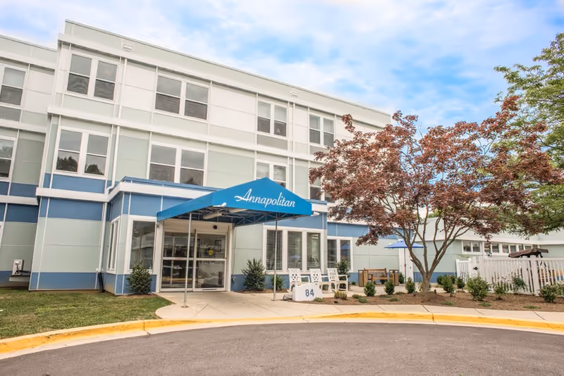 Exterior view of Annapolitan Care Center showing a three-story building with a blue and white facade. There is a blue awning above the entrance with the word 'Annapolitan' written on it. A tree with reddish leaves and some outdoor seating are visible near the entrance.
