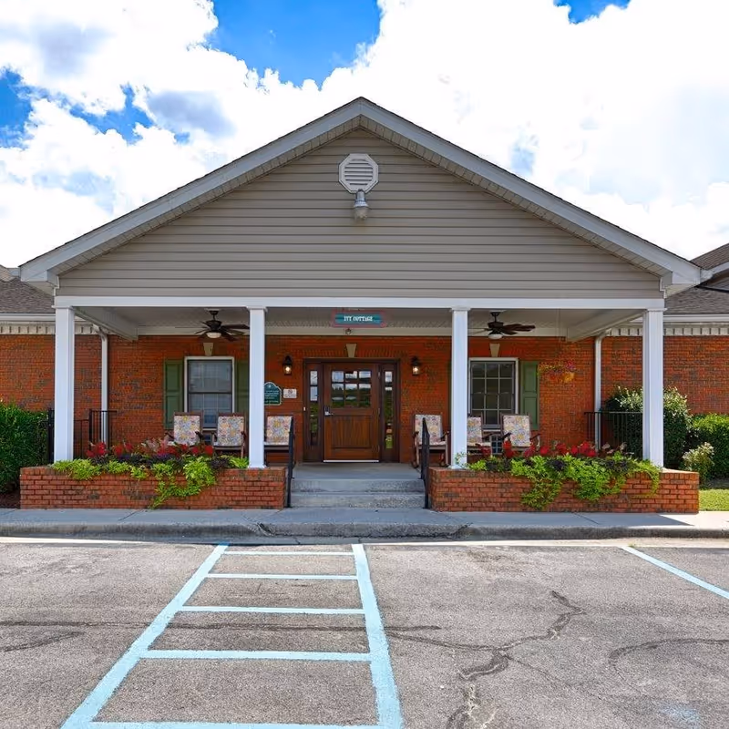 Front exterior view of a single-story brick building with a covered porch supported by white columns. The porch has ceiling fans, several chairs with floral cushions, and flower beds with green plants and red flowers. The sky is partly cloudy and there is a parking area with marked spaces in front.