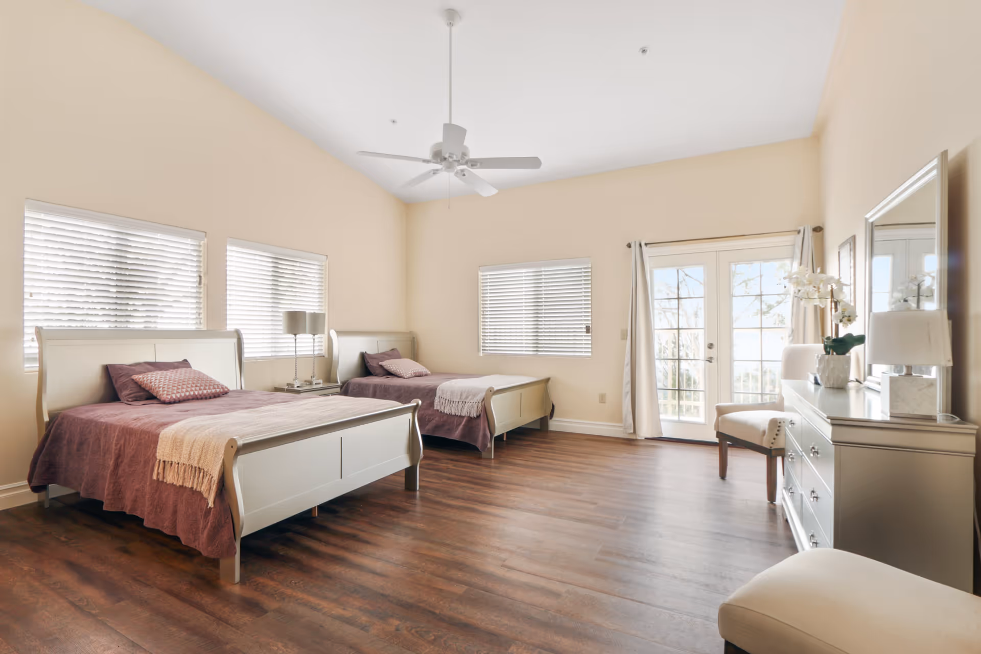 A bright bedroom with two single beds featuring white wooden frames and purple bedding. The room has wooden flooring, three windows with white blinds, a ceiling fan, a dresser with a mirror, a chair, and French doors leading outside. The walls are painted a light beige color.