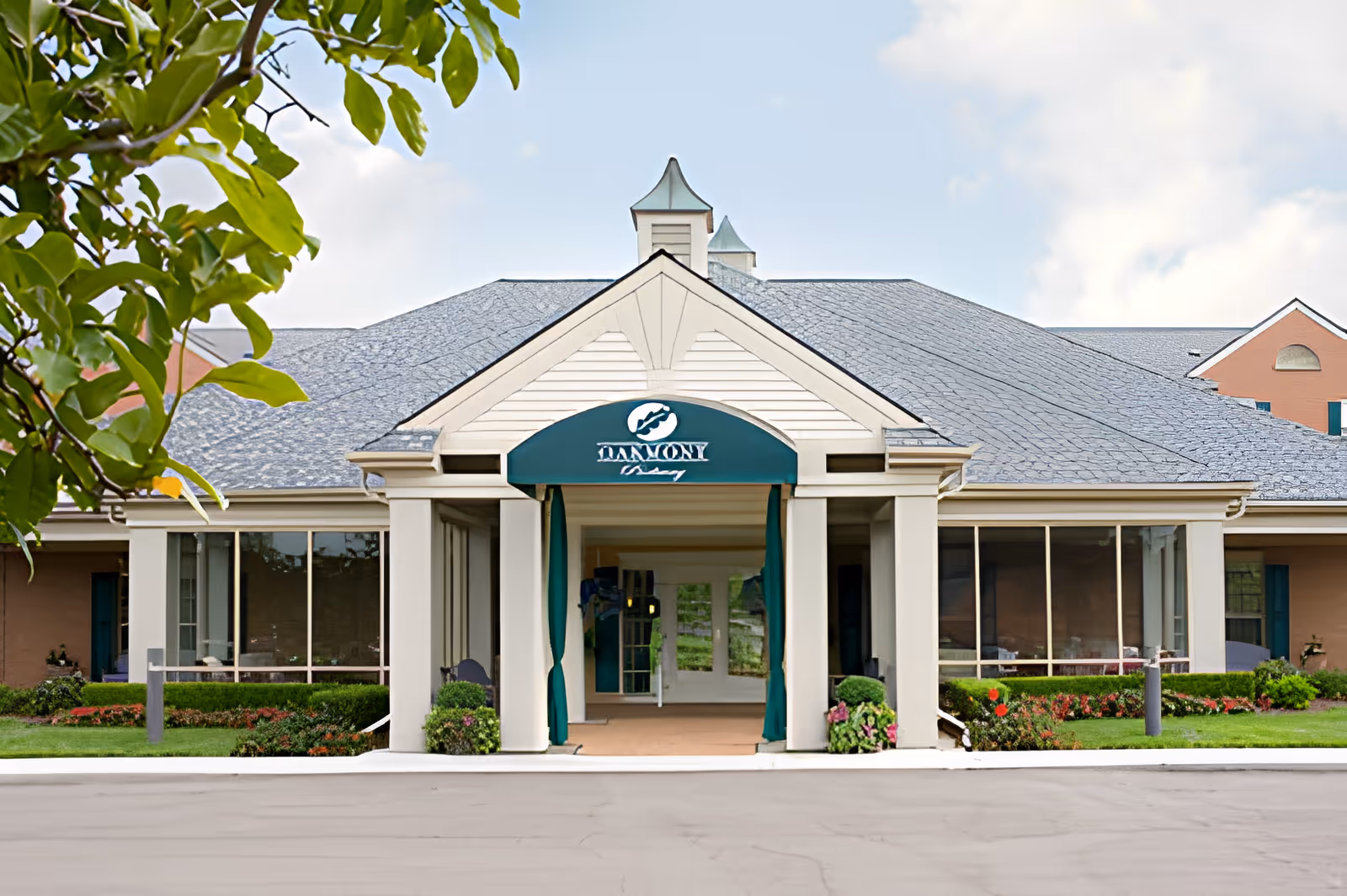 Front entrance of a single-story senior living community building with a covered portico, green awning, columns, and landscaped flower beds.