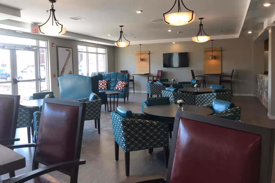 A spacious and well-lit common area with multiple round tables surrounded by patterned blue chairs and a few red leather chairs. The room features large windows allowing natural light, modern ceiling lights, a wall-mounted TV, and decorative wall panels with sconces. The floor is wooden, and the overall ambiance is welcoming and comfortable.