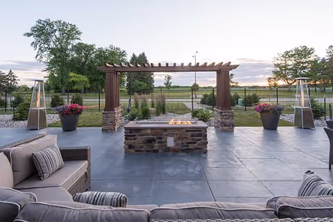 Outdoor patio area with cushioned seating around a stone fire pit, a wooden pergola, potted plants with flowers, and tall outdoor heaters, overlooking a fenced grassy area with trees under a cloudy sky at sunset.