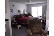 Bedroom with a red-covered bed, a cushioned armchair in the foreground, nightstands, framed pictures on the wall, and a window letting in daylight.