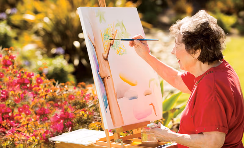 An elderly woman wearing a red shirt is painting on a canvas set on an easel outdoors, surrounded by colorful flowers and greenery in a garden setting.