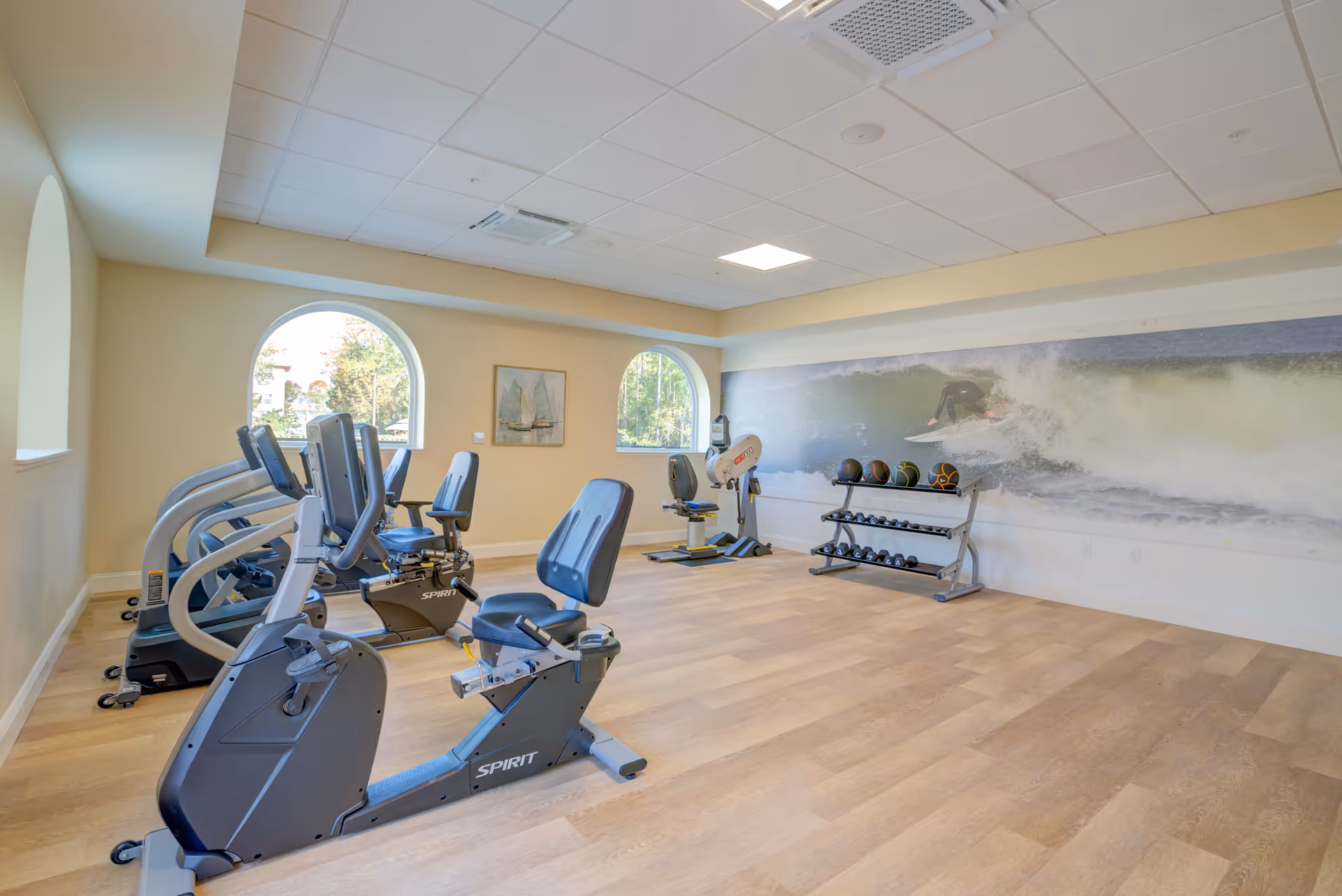Bright fitness room with recumbent exercise bikes, a weight rack, and a large ocean wave mural on the wall.