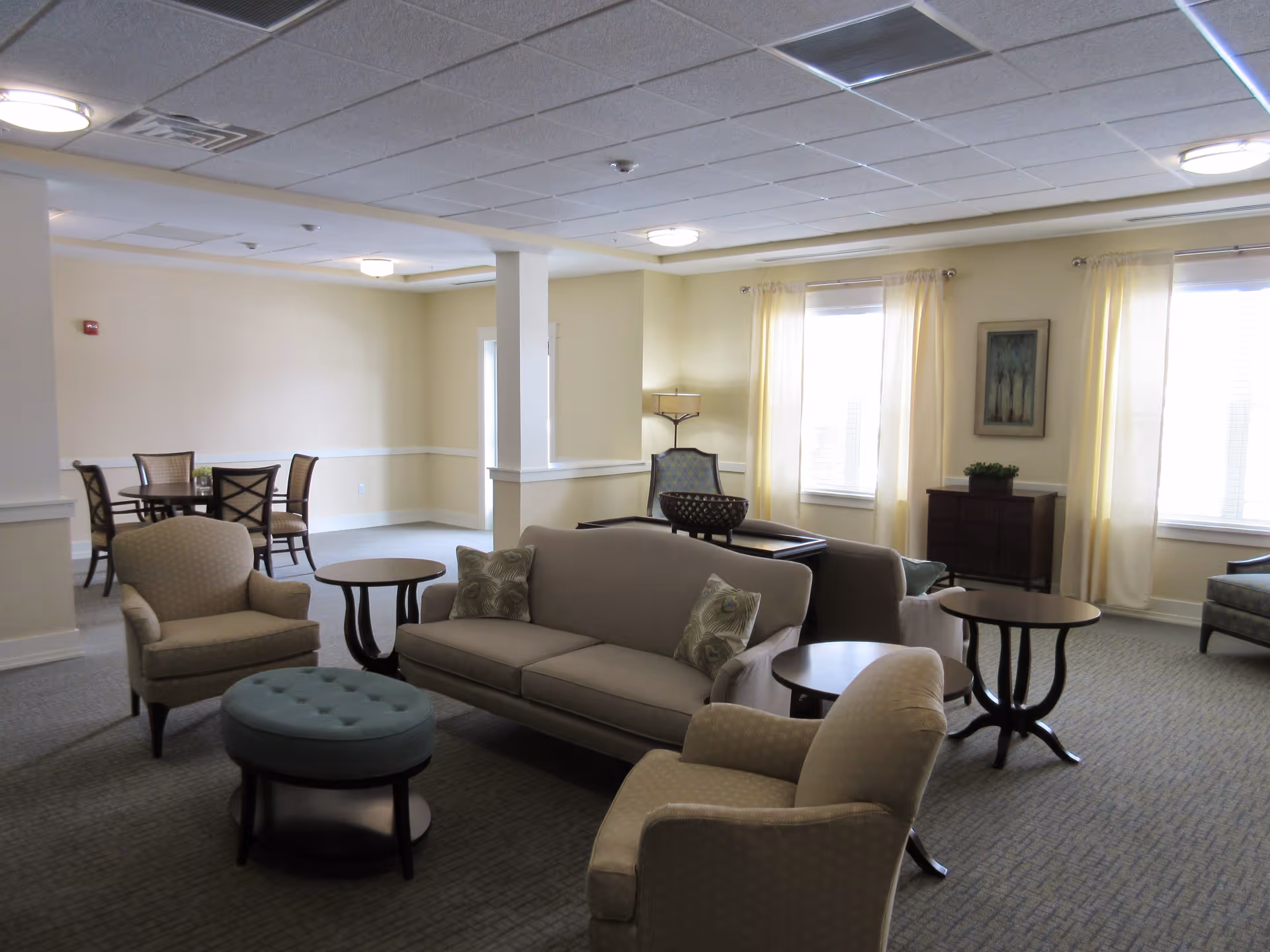 A spacious, well-lit living room area with beige walls and carpeted floor. The room features a beige sofa with patterned pillows, two beige armchairs, a blue round ottoman, and several dark wood side tables. In the background, there is a dining area with a round table and four chairs. Large windows with light curtains allow natural light to fill the room. A framed artwork and a small cabinet with a plant are visible on the far wall.