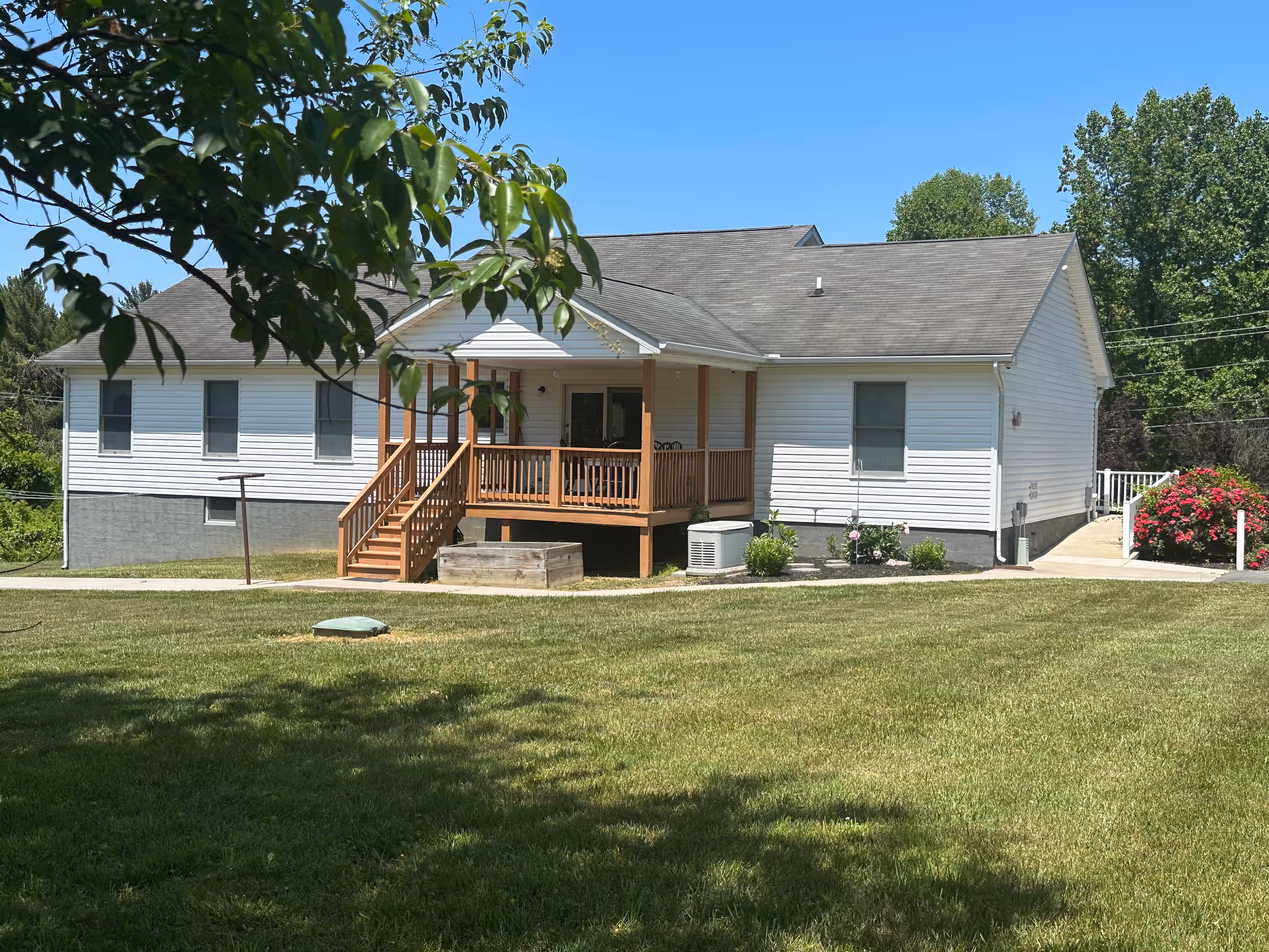 A single-story white building with a gray roof and a wooden porch with stairs leading down to a grassy lawn. The building is surrounded by trees and bushes, with a clear blue sky overhead.
